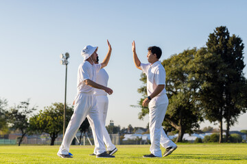 Diverse male teammates celebrating by high-fiving on suburban cricket ground wearing white uniforms