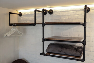 Open wall-mounted industrial wardrobe with black pipe frame and wooden shelves, white hangers, and...