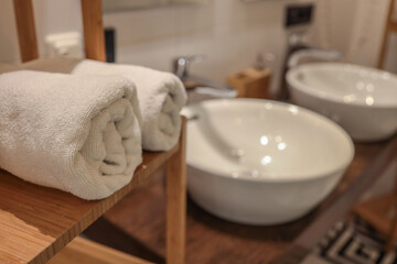 Close-up of rolled white towels on wooden shelf next to modern ceramic sinks and chrome faucets in...