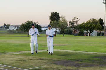 Diverse male teammates walking across cricket pitch wearing batting pads and holding wooden bats