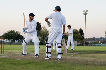 Diverse male teammates playing cricket on suburban sports ground wearing white kit and holding bats