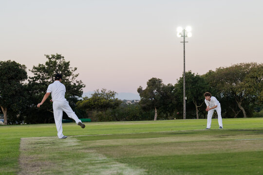 Diverse male players in cricket uniforms bowling ball toward fielder on pitch at dusk, copy space