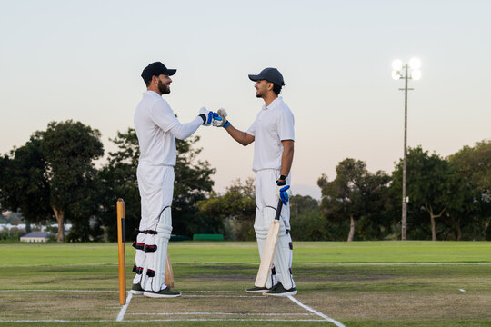 Diverse male teammates performing fist bump on marked pitch at cricket field with bats and stumps