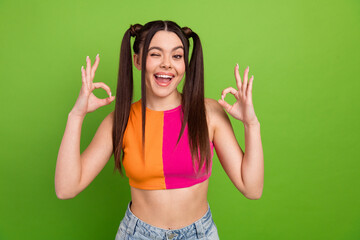 Cheerful young woman in a colorful outfit gesturing OK with both hands against a vibrant green...