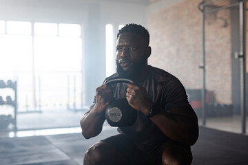 African American man holding 16 kg kettlebell, squatting on rubber floor in gym with dumbbell rack