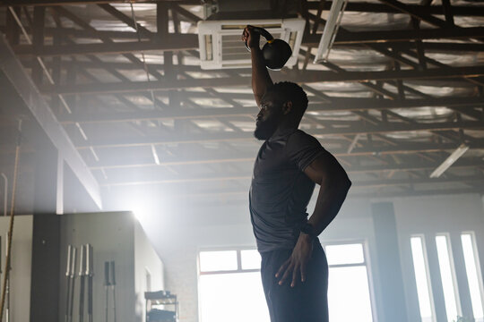 Athlete pressing kettlebell overhead in gym with exposed beams and AC, barbell rack, rope, windows