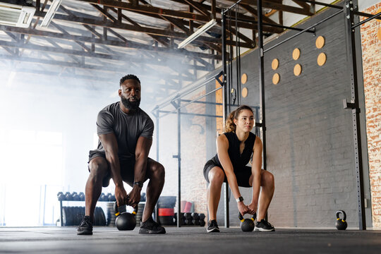 Diverse training partners wearing sportswear lifting kettlebells in squat stance in CrossFit gym