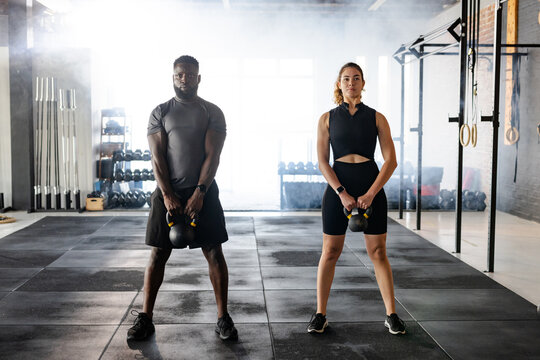 Diverse workout partners holding kettlebells on rubber mat floor at modern gym with pull-up rig