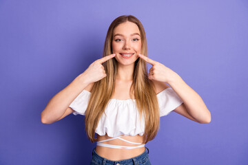 Confident young woman wearing braces pointing at her smile, posing on purple background,...