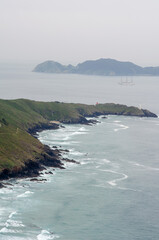 Scenic view of rugged green cliffs meeting the Atlantic Ocean, with a tall ship sailing in the distance under a cloudy sky. Ideal for travel and nature concepts.