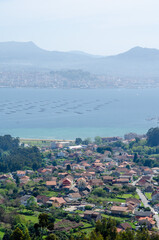 Scenic view of a picturesque Galician village by the sea with distant mountains and mussel farms, capturing peaceful rural life and beautiful spring landscape.