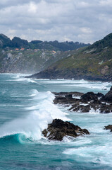 Powerful turquoise waves break against rugged cliffs on a remote coastline, with distant houses and lush green hills under moody clouds creating a dramatic atmosphere.