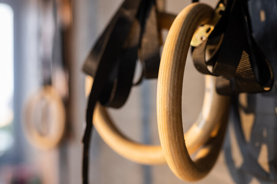 Wooden gymnastic ring hanging on black strap against grey wall near window with blurred ring behind