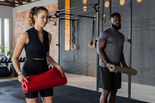 Diverse workout partners holding yoga mats and stretching in fitness studio near pull-up rig - Powered by Adobe