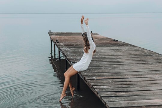 unrecognizable woman in a white dress, sitting on the edge of wooden pier, dipping her feet into calm water with a light splash, gazing at horizon under soft cloudy sky, relaxation, travel