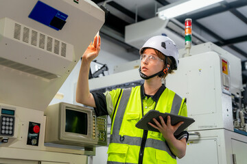 A woman wearing a yellow vest and a hard hat is looking at a machine. Female engineer working with automated machinery in a smart factory, concept of Industry 4.0 and manufacturing technology.