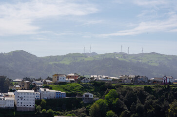 Peaceful rural village with colorful houses on lush green hills, distant wind turbines, and clear blue sky, capturing sustainable energy and serene countryside life.