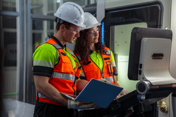 Two people wearing orange vests and hard hats are looking at a laptop computer. Team of engineers programming and operating a CNC machine in a smart factory.
