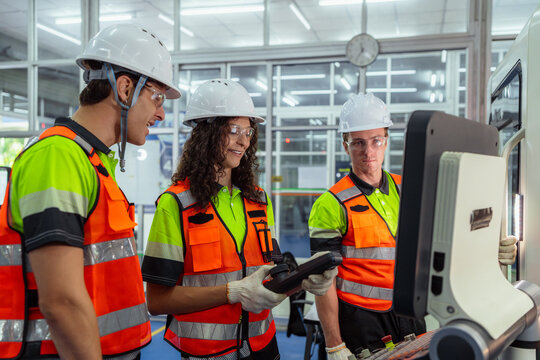 Three people wearing safety gear are standing in front of a computer monitor. Female instructor training new workers or apprentices on how to operate a CNC machine in a factory.