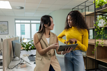 Asian Chinese and African American female coworkers standing at office desks discussing tablet