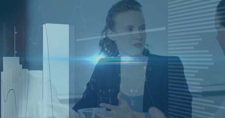 Presenting woman in blazer gesturing toward colleague at conference table, with financial charts