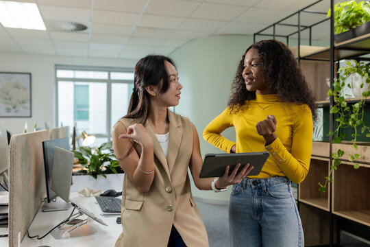 Diverse female coworkers discussing strategy on tablet beside white desks in open-plan office