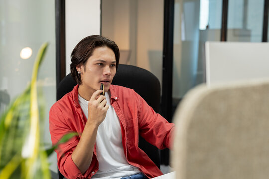 Asian man sitting at desk in modern office holding pen near chin gazing at large monitor