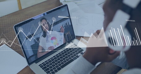 Listening man wearing over-ear headphones viewing chart on laptop at desk with notebook, papers
