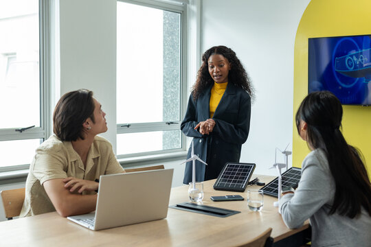 Diverse coworkers discussing renewable energy using laptop, turbines and solar panels at office - Powered by Adobe