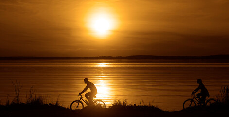 sporty couple friends on bicycles outdoors against sunset. Silhouette of motion go of two 2 cyclist along shoreline coast Reflection sun on water. Empty Copy Space for inscription, people or objects.