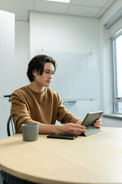 Asian man wearing brown sweater scrolling tablet at office with whiteboard smartphone and grey mug