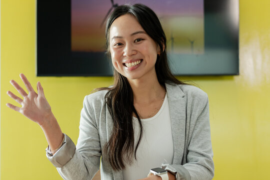 Asian woman smiling and waving in conference room at desk by widescreen display showing turbines
