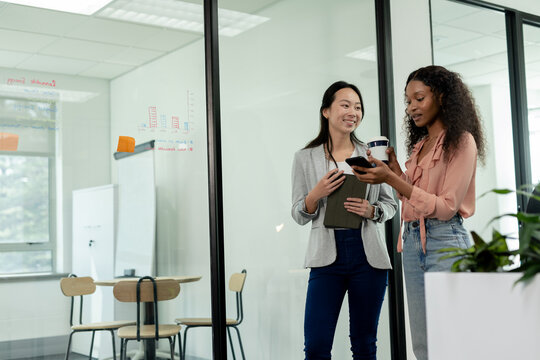 Diverse female colleagues holding smartphone cup and tablet by glass wall in office, copy space