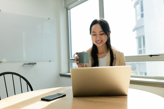 Asian woman holding coffee mug and smiling at laptop on office table with smartphone and whiteboard - Powered by Adobe