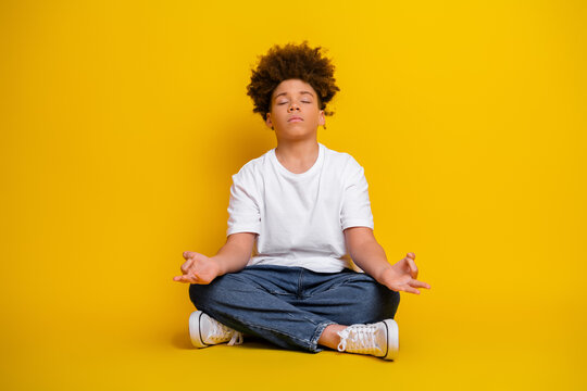 Young boy practicing meditation while sitting on floor against a yellow background. The child is in focus with a calm demeanor.