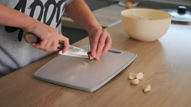 Close-up of a woman hands chopping fresh garlic with a large kitchen knife on a scratched cutting board. 