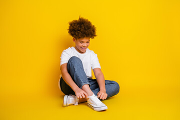 Happy young boy sitting cross-legged on vibrant yellow background, tying his white sneakers and...