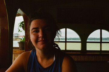 A woman seated in a cozy oceanside interior with natural lighting, looking relaxed and thoughtful against the backdrop of the calm ocean seen through large windows adorned with curved frames.35mm film