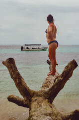 A woman on a tropical beach stands on a driftwood log overlooking serene turquoise waters, with a boat anchored in the distance, evoking a sense of adventure, relaxation, and scenic beauty. 35mm film