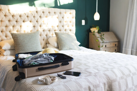Open beige suitcase resting on made bed with patterned pillows and headphones against teal wall