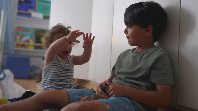 Baby flinches and laughs nervously as older brother dangles plush keychain above head, both children seated on floor in playroom surrounded by books and toys
