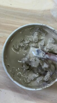 Close-up of hands mixing clay with water in a ceramic bowl to make slip. Artistic ceramic process in natural light, surrounded by wildflowers and rustic tools on a wooden table
