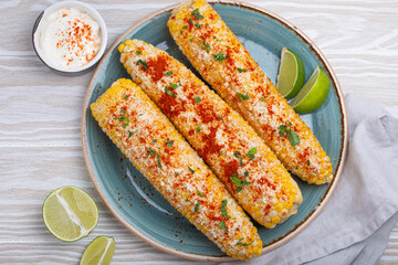 Three grilled corn cobs coated in spices, mayonnaise, and cotija cheese are served on a plate with...