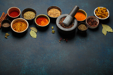 Colorful bowls of aromatic spices and herbs on a dark background, with a mortar and pestle,...