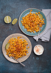 Two plates of esquites, a popular mexican street food dish, are served with lime wedges and sour cream with chili powder