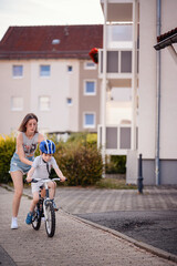 Mother Helping Son Learn to Ride a Bike on Sidewalk