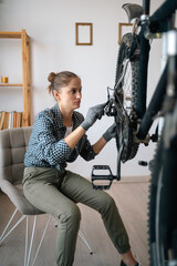 woman sitting on chair repairing bicycle by herself at home adjusting chain and fixing rear wheel,...