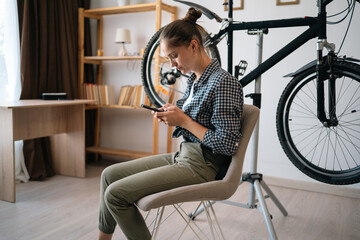 woman sitting on chair at home repairing bicycle at home reading online tips using AI assistant,...