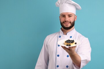 Happy confectioner in uniform holding tart of blueberries on light blue background