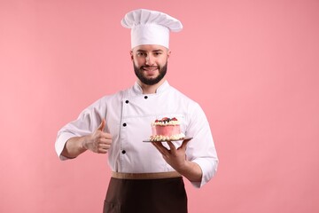 Happy confectioner in uniform holding cake with berries and showing thumbs up on pink background